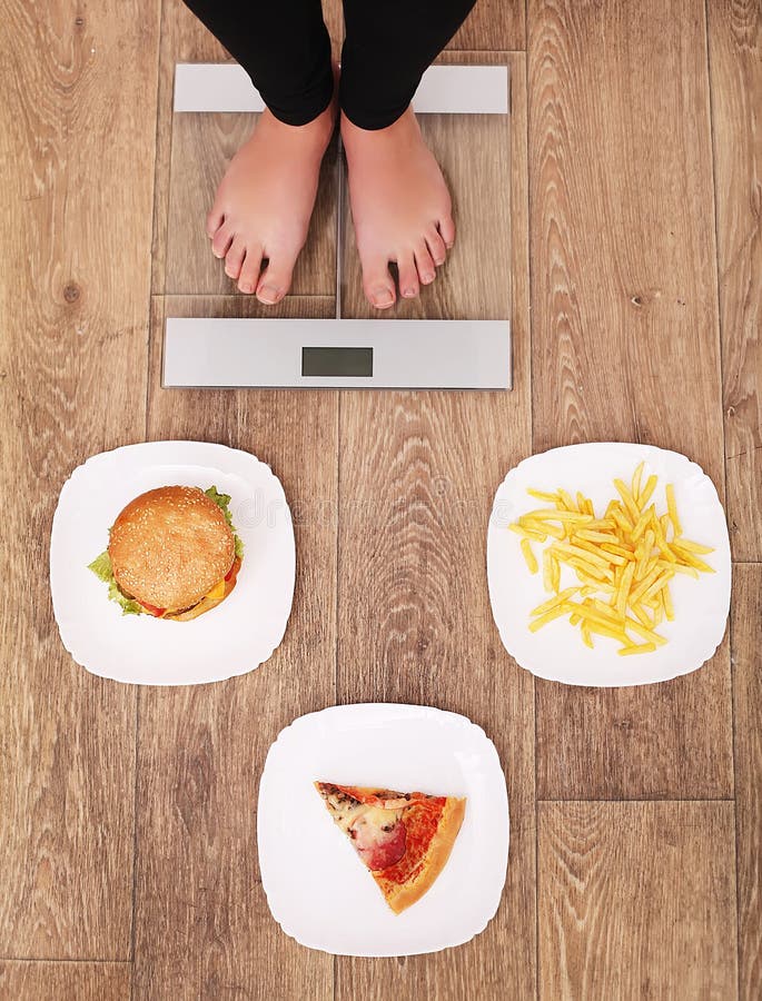 Una mujer hermosa joven está en las escalas Para elegir entre las patatas fritas, la pizza y las hamburguesas El concepto de cons fotografía de archivo Una mujer hermosa joven está en las escalas Para elegir entre las patatas fritas, la pizza y las hamburguesas El concepto de cons fotografía de archivo
