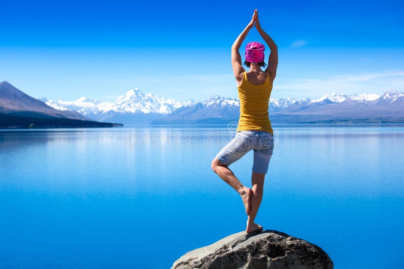 Una mujer joven atractiva que hace una actitud de la yoga para la balanza y que estira cerca del lago