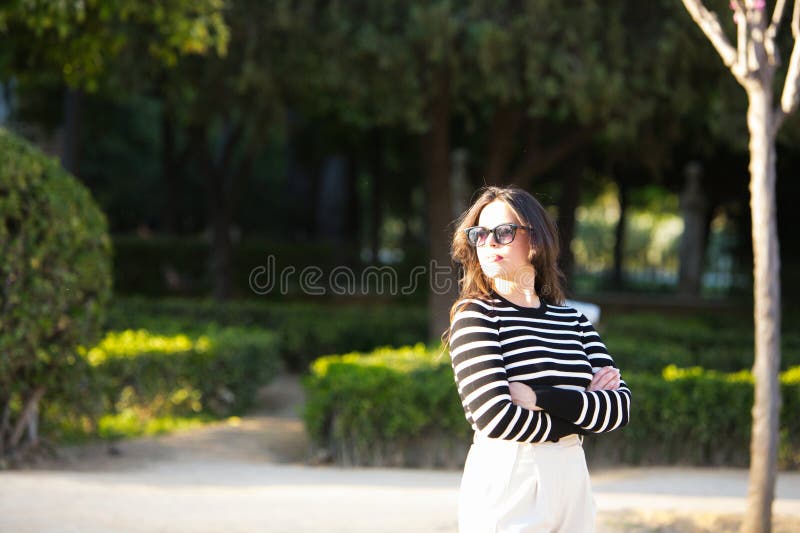 Una mujer joven y bonita con un largo pelo castaño vestido con una camisa de rayas blancas y negras y pantalones blancos y brazos fotografía de archivo libre de regalías