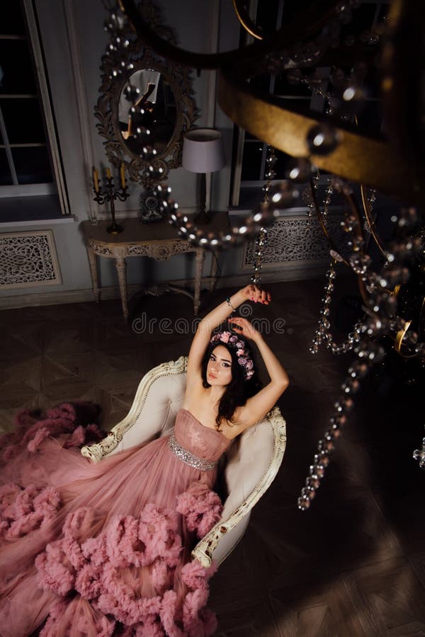 Una mujer morena delgada hermosa en un vestido rosado con volantes se está sentando en una butaca azul del terciopelo imagenes de archivo Una mujer morena delgada hermosa en un vestido rosado con volantes se está sentando en una butaca azul del terciopelo imagenes de archivo
