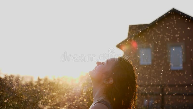 Una mujer sonriente feliz está bajo la lluvia. impresionantes rayos solares dorados brillan en una joven lúdica disfrutando de una almacen de metraje de vídeo