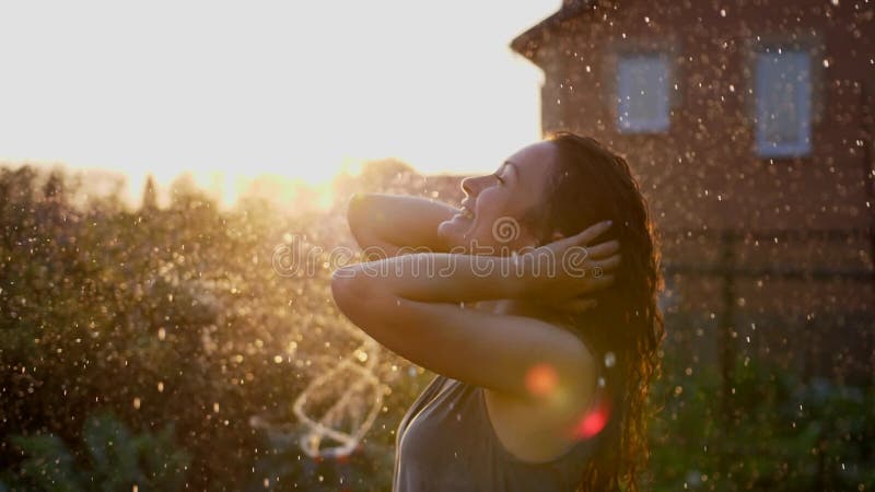 Una mujer sonriente feliz está bajo la lluvia. impresionantes rayos solares dorados brillan en una joven lúdica disfrutando de una almacen de metraje de vídeo