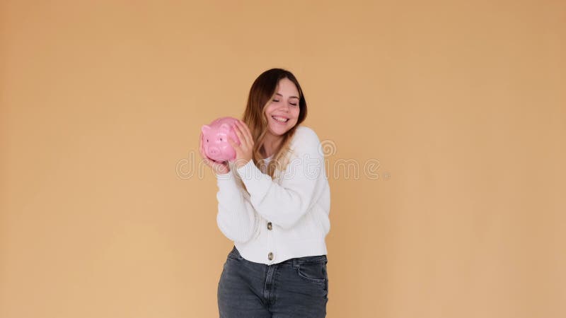 Una mujer sonriente sosteniendo un banco de cerdo y poniendo una moneda en el fondo beige almacen de metraje de vídeo