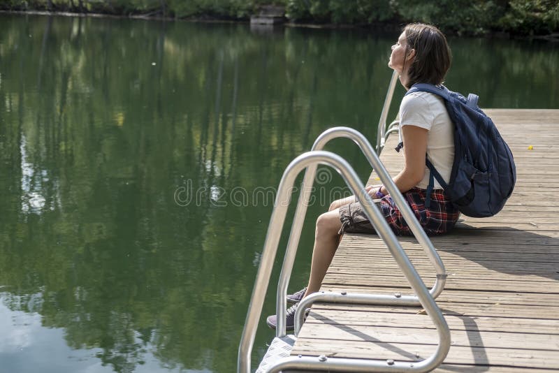 Una Mujer Tranquila Y SoÃ±adora Con Mochila Se Sienta En Muelle De Madera Junto Al Lago, En Los Rayos Del CÃ¡lido Sol De Verano de archivo - Imagen de embarcadero,