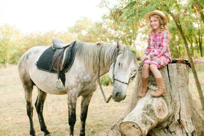 A nice little girl with light curly hair in a vintage plaid dress and a straw hat and a gray horse. Rural life in autumn. Horses and people. A nice little girl with light curly hair in a vintage plaid dress and a straw hat and a gray horse. Rural life in autumn. Horses and people