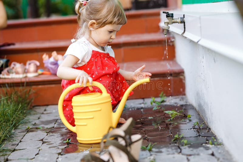 Una niña vestida en los sundress está dibujando el agua en una regadera en el jardín al lado de la casa en el día de verano imagen de archivo Una niña vestida en los sundress está dibujando el agua en una regadera en el jardín al lado de la casa en el día de verano imagen de archivo