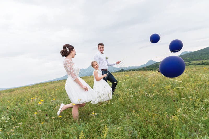 Una pareja joven y su hija en vestidos de boda están caminando en naturaleza con los globos imagen de archivo