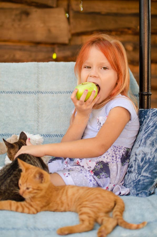 Una pequeña muchacha caucásica de seis años se está sentando en un oscilación del jardín, está jugando con los gatitos y está com imágenes de archivo libres de regalías Una pequeña muchacha caucásica de seis años se está sentando en un oscilación del jardín, está jugando con los gatitos y está com imágenes de archivo libres de regalías