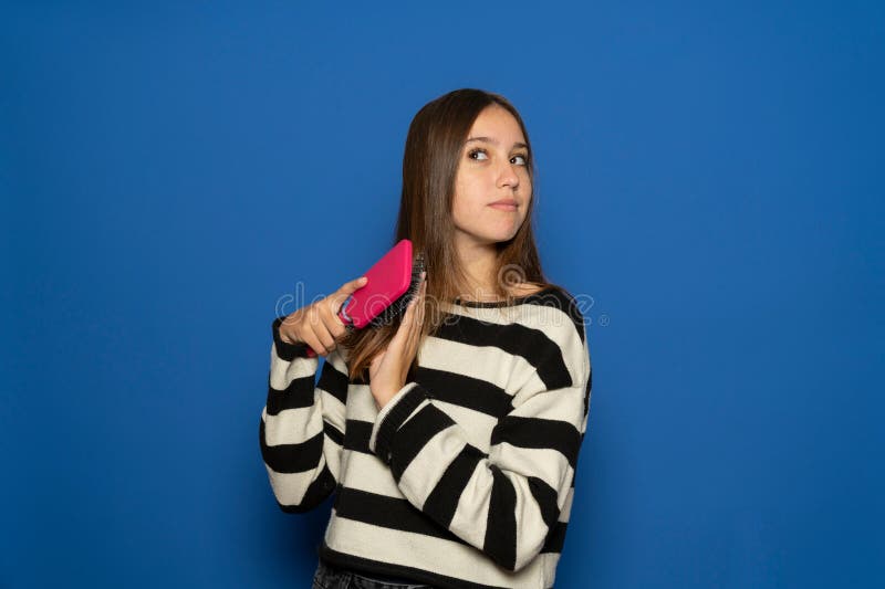 Una ragazza ispanica con un maglione striato felice di essersi pettinata i capelli di seta con una spazzola in piedi sopra il blu fotografia stock