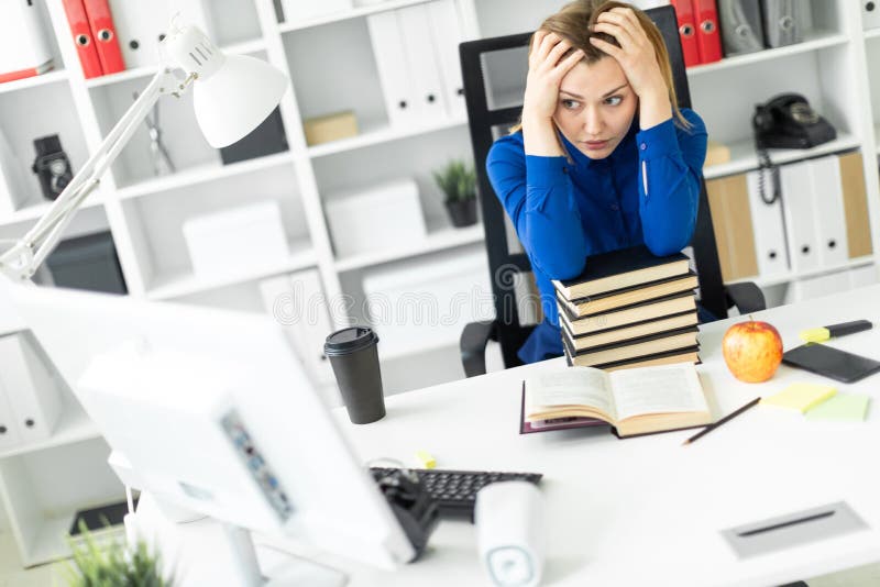 Una ragazza si siede ad uno scrittorio del computer e tiene le sue mani dietro la sua testa Prima della ragazza si trova un libro. La bella ragazza in una camicia blu sta lavorando in un ufficio luminoso Foto con profondità di campo