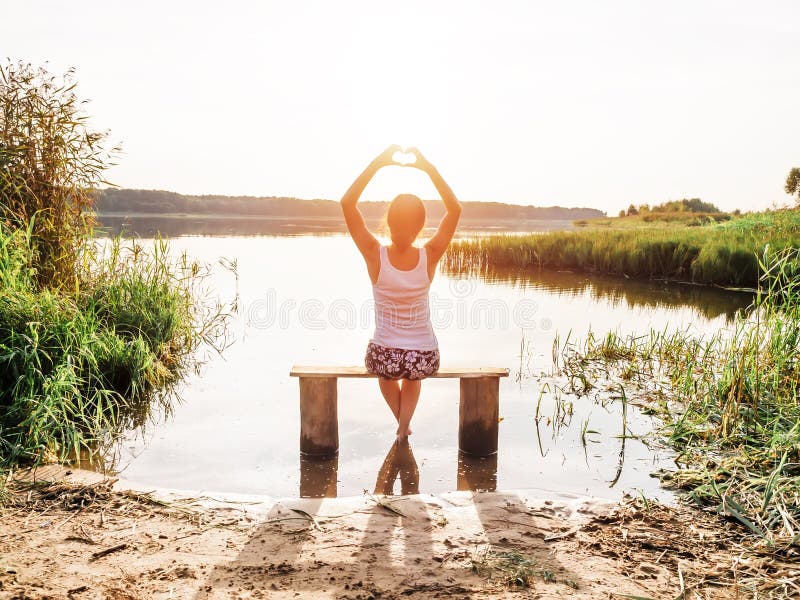 Una ragazza si siede su un banco dal fiume gode di bello tramonto e mostra il segno del cuore delle dita delle mani Ragazza nella fotografia stock Una ragazza si siede su un banco dal fiume gode di bello tramonto e mostra il segno del cuore delle dita delle mani Ragazza nella fotografia stock