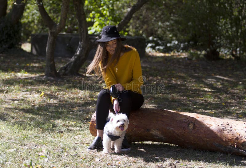 Una ragazza sorridente che si siede in un parco che guarda un poco ddog bianco immagine stock Una ragazza sorridente che si siede in un parco che guarda un poco ddog bianco immagine stock
