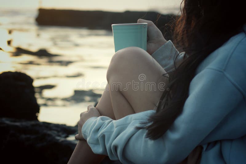 Young woman sitting on sea rock, holding a cup of morning tea or coffee in the beach. Self care concept. Relax on weekend or on vacation concept. Young woman sitting on sea rock, holding a cup of morning tea or coffee in the beach. Self care concept. Relax on weekend or on vacation concept.