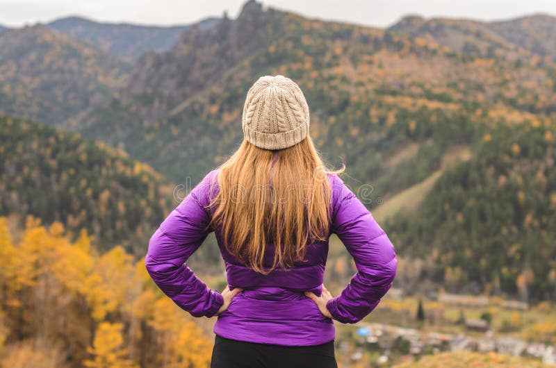 Una ragazza in un rivestimento lilla guarda fuori nella distanza su una montagna, in una vista delle montagne ed in una foresta a fotografia stock Una ragazza in un rivestimento lilla guarda fuori nella distanza su una montagna, in una vista delle montagne ed in una foresta a fotografia stock