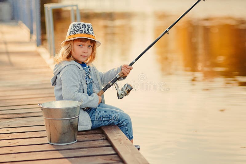 Una ragazzina pescatrice sorride seduta vicino a un fiume con una canna fotografia stock Una ragazzina pescatrice sorride seduta vicino a un fiume con una canna fotografia stock