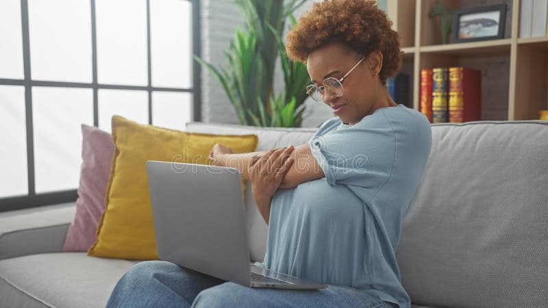 Una relajada mujer afroamericana que estira su brazo durante un descanso por usar su laptop en un moderno salón foto de archivo libre de regalías