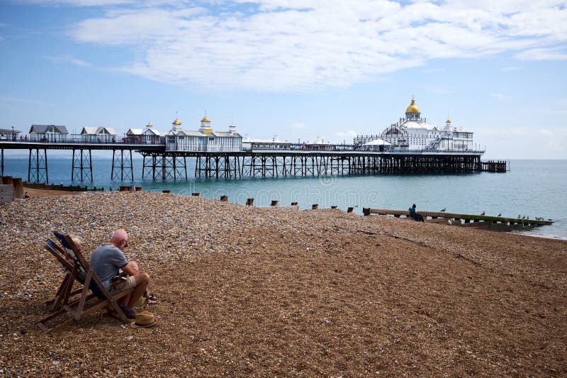 Eastbourne, East Sussex | UK - 4 August 2021: A middle-aged sitting in deckchairs on the beach with landmark Victorian pier in the background. Eastbourne, East Sussex | UK - 4 August 2021: A middle-aged sitting in deckchairs on the beach with landmark Victorian pier in the background