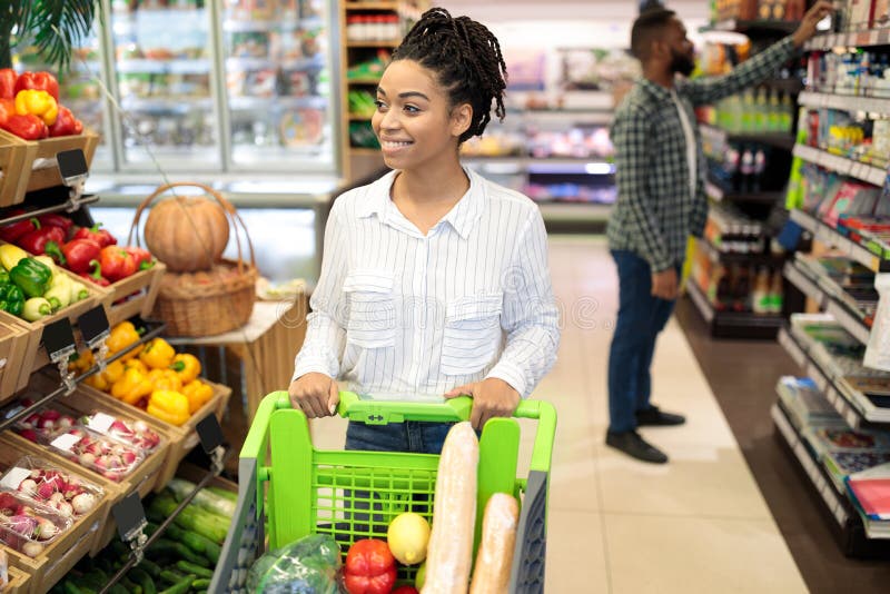 African American Lady Buying Fresh Organic Fruits And Vegetables Doing Grocery Shopping Walking With Shop Cart In Supermarket Indoors. Customer Of Groceries Store Choosing Healthy Natural Food. African American Lady Buying Fresh Organic Fruits And Vegetables Doing Grocery Shopping Walking With Shop Cart In Supermarket Indoors. Customer Of Groceries Store Choosing Healthy Natural Food