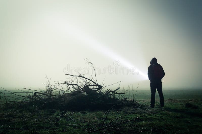 Una siluetta sinistra di una figura incappucciata sola in un campo su una notte nebbiosa con una torcia Con un buio pubblichi