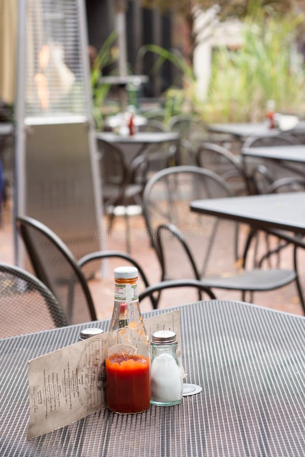 A metal outdoor restaurant table with a ketchup bottle, salt shaker, and menu. An outdoor heater and other tables are in the background. A metal outdoor restaurant table with a ketchup bottle, salt shaker, and menu. An outdoor heater and other tables are in the background.