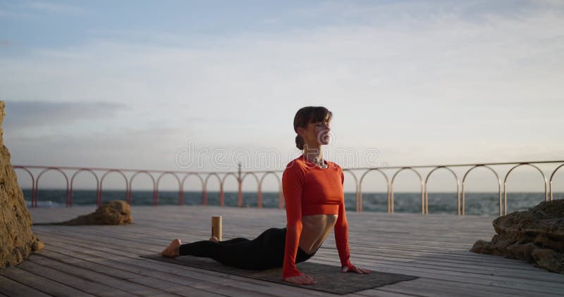 Una valiente morena amante del yoga, con un suéter rojo en una alfombra de yoga, propone una cobra por la mañana cerca del mar almacen de metraje de vídeo