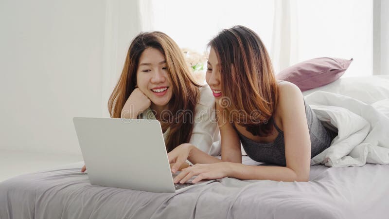 Underbara unga asiatiska kvinnor, glada lesbiska par som sitter på sängen och använder datorn tillsammans hemma i sovrummet. lgbt fotografering för bildbyråer