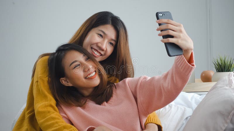 Underbara unga asiatiska kvinnor, lesbiskt lyckligt par som sitter på soffhug och använder telefon för att ta selfig tillsammans i royaltyfri fotografi