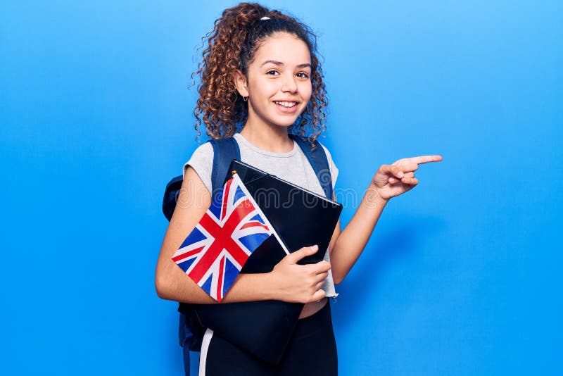 Beautiful kid girl with curly hair wearing student backpack holding binder and uk flag smiling happy pointing with hand and finger to the side. Beautiful kid girl with curly hair wearing student backpack holding binder and uk flag smiling happy pointing with hand and finger to the side