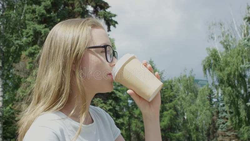 Underbart blondinskt leende hona i glasögon som ser fromt ut från kameran och dricker kaffe för att gå och stå i