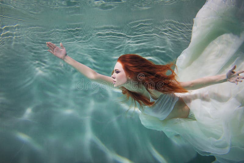 Underwater girl. Beautiful red-haired woman in a white dress, swimming under water.