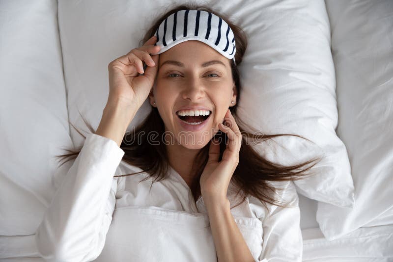 Happy healthy beautiful young woman wear sleeping mask awake after sleep laughing lying in comfortable bed on pillow, smiling lady waking up enjoy good morning looking at camera, portrait top view. Happy healthy beautiful young woman wear sleeping mask awake after sleep laughing lying in comfortable bed on pillow, smiling lady waking up enjoy good morning looking at camera, portrait top view