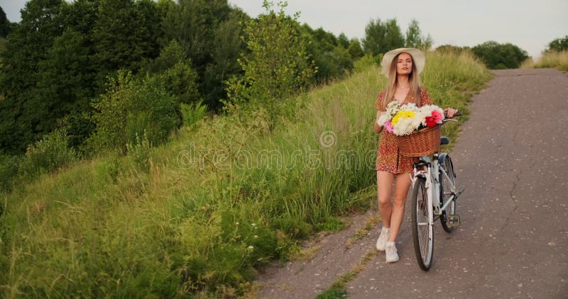 Une belle femme avec une bicyclette dans un chapeau et une robe légère d'été vient avec des fleurs dans un panier et des sourires