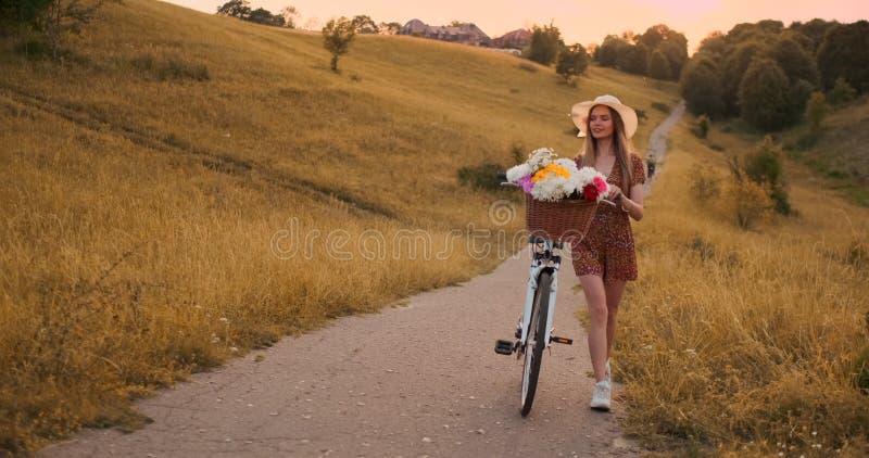 Une belle femme avec une bicyclette dans un chapeau et une robe légère d'été vient avec des fleurs dans un panier et des sourires