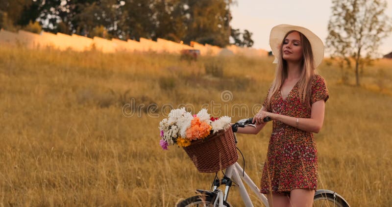 Une belle femme avec une bicyclette dans un chapeau et une robe légère d'été vient avec des fleurs dans un panier et des sourires