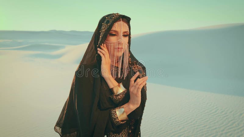 Une belle femme en robe orientale noire se dresse dans le désert. Modèle de mode posant. La tête est recouverte d'un banque de vidéos
