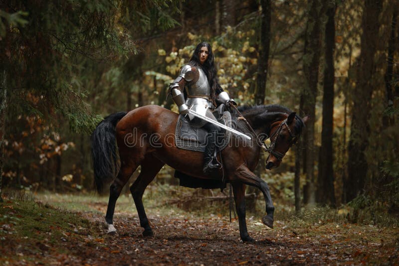 Une belle fille de guerrier avec un chainmail et une armure de port d'épée montant un cheval dans une forêt mystérieuse images stock Une belle fille de guerrier avec un chainmail et une armure de port d'épée montant un cheval dans une forêt mystérieuse images stock