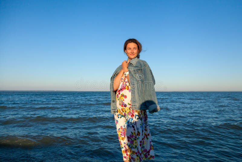 Une brune gaie dans la veste colorée de fil et de denim sourit, marche le long de la plage et apprécie le soleil lumineux sur a photos stock Une brune gaie dans la veste colorée de fil et de denim sourit, marche le long de la plage et apprécie le soleil lumineux sur a photos stock