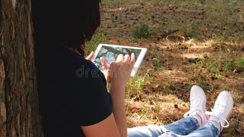 Une brunette inconnue assise dans un arbre du parc et travaillant avec un ordinateur à tablettes. Tourisme féminin utilisation d banque de vidéos