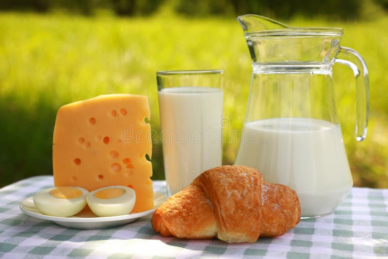 Une cruche de lait, un verre de lait, un morceau de fromage et d'un oeuf coupé d'un plat, et un croissant sur une nappe à carreau photo stock