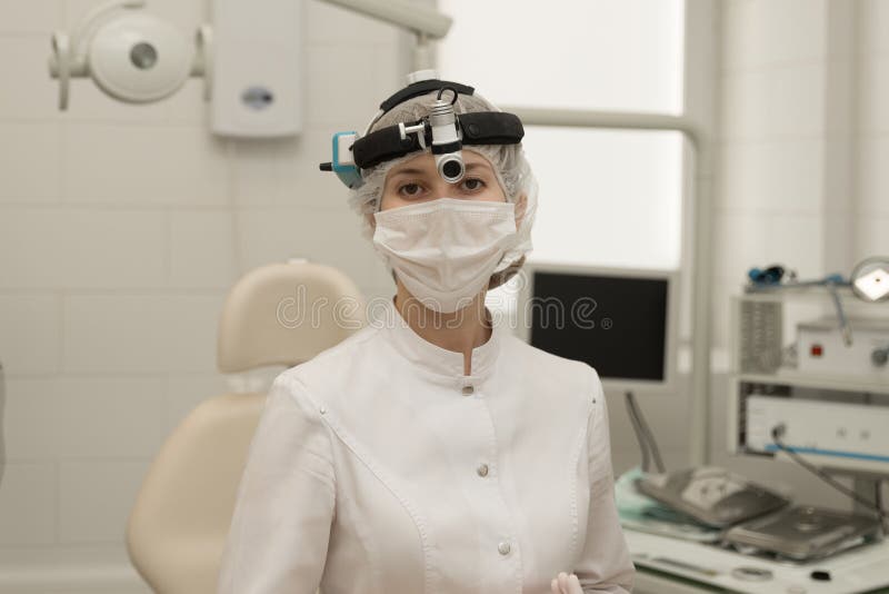 Une doctorotolaryngologiste dans un bureau moderne de l'hôpital regarde dans la caméra dans un manteau blanc et un masque