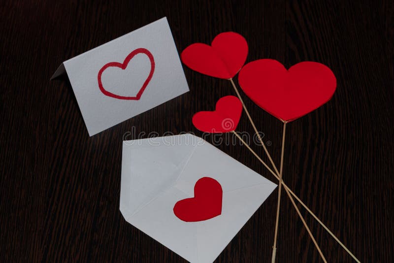 A white envelope with a red heart,a love message and flags in the form of red cardboard hearts. Attributes of Valentine's day,on a brown,textured background,with a wood texture. A white envelope with a red heart,a love message and flags in the form of red cardboard hearts. Attributes of Valentine's day,on a brown,textured background,with a wood texture.