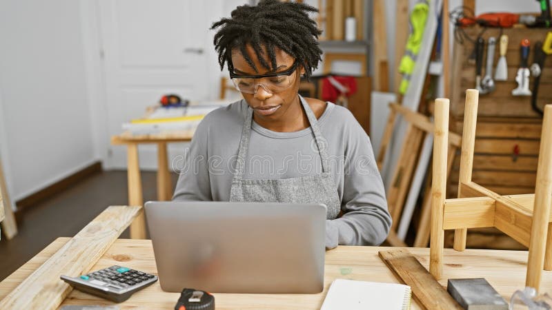 Une femme afro-américaine avec des dreadlocks travaillant sur un ordinateur portable dans un atelier de travail du bois image libre de droits