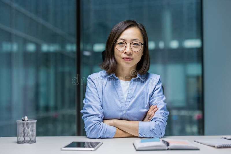 Une femme asiatique confiante avec des lunettes est assise à un bureau bras croisé dans un environnement de bureau moderne prêt à photo libre de droits