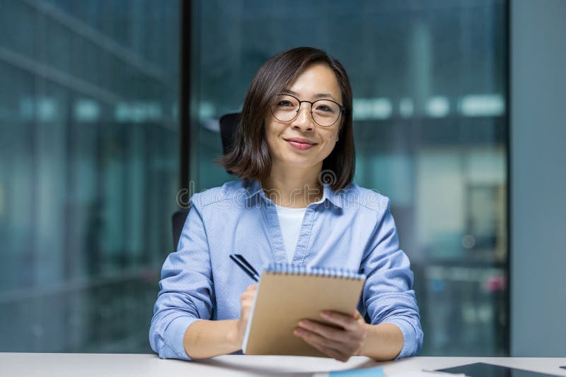 Une femme asiatique en lunettes est assise sur un bureau tenant un ordinateur portable regardant directement la caméra avec un sou images stock