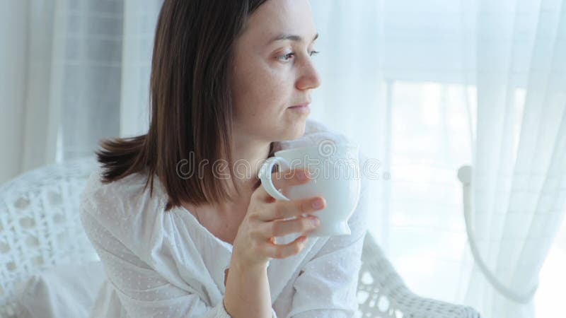 Une femme assise dans un fauteuil prend une tasse et des boissons en regardant par la fenêtre