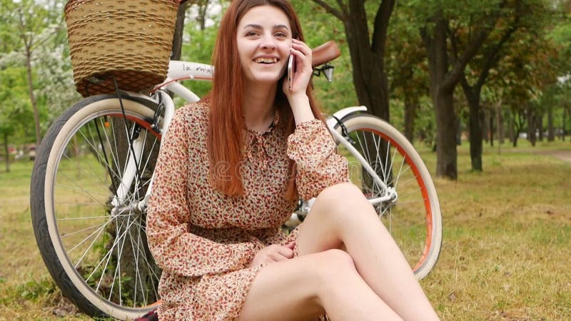 Une femme assise sur l'herbe dans un parc de la ville. location de vélo repos extérieur. la fille utilise son téléphone pour commu banque de vidéos