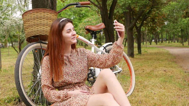 Une femme assise sur l'herbe dans un parc de la ville. location de vélo repos extérieur. la fille utilise son téléphone pour commu banque de vidéos