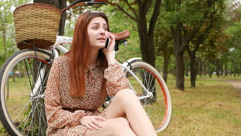 Une femme assise sur l'herbe dans un parc de la ville. location de vélo repos extérieur. la fille utilise son téléphone pour commu banque de vidéos
