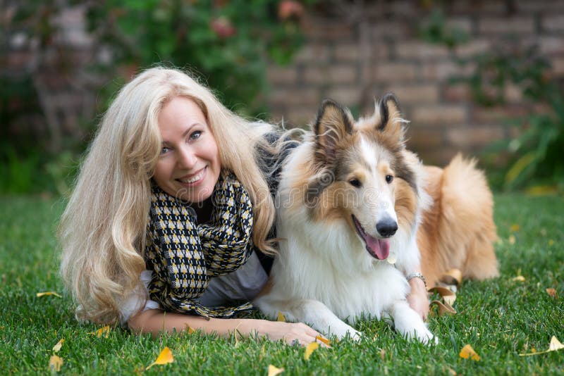 Une femme blonde caucasienne allongée sur une herbe verte serrant son collie photographie stock Une femme blonde caucasienne allongée sur une herbe verte serrant son collie photographie stock