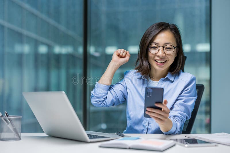 Une femme d'affaires asiatique enthousiaste célèbre son succès en regardant son téléphone quand elle est à son bureau. photo stock